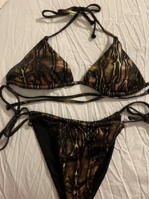 NY! Svart och guld orm bikini - NY! Snygg vadderad trekantsbikini med svart och guldigt ormmönster. Både överdel och underdel har knytband för justerbar passform. 