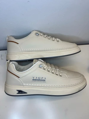 Vita sneakers med textdetalj - Stilrena vita sneakers från Y3QPA med låg profil och textdetalj på sidan. Skorna har vita snören, grå sula och en brun detalj vid hälen. Tillverkade i slätt syntetmaterial med modern design och diskreta loggor på sidan och sulan. Helt nya utan kvitto eller orginalförpacknig 