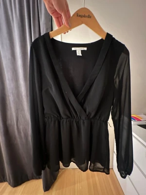 Svart omlottblus från Nelly - Snygg svart omlottblus från Nelly med transparenta långärmar och markerad midja. Blusen har djup v-ringning och är gjord i ett lätt, tunt material som ger ett elegant fall. Perfekt att styla med jeans eller kjol för en trendig look.