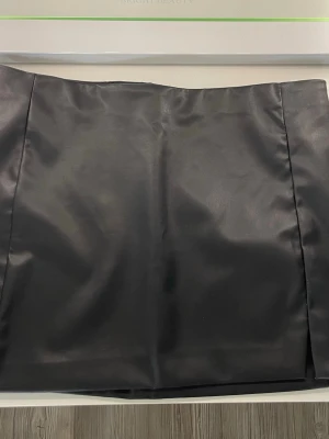 Svart pennkjol i fuskläder - Säljer en svart pennkjol i fuskläder med slät yta och enkel design. Tjolen är använd en gång och är i mycket bra skick. 