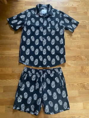 Svart set med vita palmbladsmönster - Snyggt och somrigt set! Kortärmad skjorta och shorts i svart med vita palmbladsmönster. Skjortan har klassisk krage och knäppning framtill, ficka i fram, shortsen har snöre i midjan och sidofickor, samt ficka ibak. Perfekt för en avslappnad och trendig look. Skjortan är storlek M och shortsen är storlek S! 