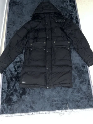 Svart dunjacka från Haglöfs med huva - Säljer en svart lång dunjacka från Haglöfs med huva och flera stora fickor framtill. Jackan har en stilren design med diskret logga på bröstet och tryckknappar. Perfekt för kalla vinterdagar när du vill hålla dig varm och bekväm.