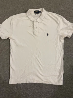 Vit pikétröja från Polo Ralph Lauren - Klassisk vit pikétröja från Polo Ralph Lauren med broderad marinblå logga på bröstet. Tröjan har krage, knappar framtill och är i en slim fit-modell. Tillverkad i bomull för en skön och luftig känsla.