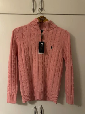 Rosa kabelstickad tröja Polo Ralph Lauren - Kabelstickad rosa tröja från Polo Ralph Lauren med halv dragkedja och ribbade muddar. Klassisk logga broderad på bröstet. Tröjan har hög krage och är tillverkad i mjukt material, perfekt för en chill och snygg look.