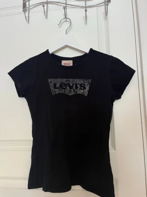 Svart Levi's t-shirt med tryck - Säljer en svart t-shirt från Levi's med ett stort logotryck i vitt och svart framtill. T-shirten har rund halsringning och korta ärmar. Materialet känns mjukt och skönt, troligtvis bomull. Perfekt till jeans eller shorts!