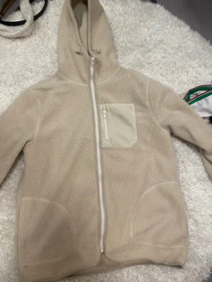 Beige teddyjacka med huva - Mysig beige teddyjacka med huva och sneddragkedja. Jackan har en ljus bröstficka med dragkedja och två stora fickor framtill. Oversize