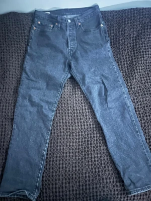 Levi's 501 jeans - Klassiska mörkgråa jeans från Levi's med rak passform och fem fickor. Jeansen har knappgylf, nitar vid fickorna och den ikoniska röda Levi's-lappen bak. Tillverkade i slitstark denim som ger en tidlös look. Storlek W27 L28 ganska nya säljer för dem är för små skriv privat för andra frågor!
