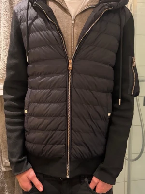 Svart Moncler pufferjacka med huva - Svart pufferjacka från Moncler med quiltad framsida, stickade ärmar och huva med stor logga. Jackan har dragkedja fram, ficka med dragkedja på ärmen och snygga detaljer i metall. Perfekt för dig som vill ha en stilren och modern look. Storlek moncler 2