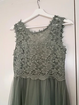 Lång grön spetsklänning - Elegant långklänning i ljusgrön nyans med vacker spets upptill och transparent tyllkjol. Ärmlös modell med rund halsringning och fina blommiga spetsdetaljer. Perfekt för speciella tillfällen när du vill känna dig extra fin.