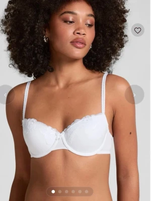 Vit BH med spets från hunkemöller  - Säljer en vit BH från hunkemöller med vacker spetsdetalj på övre delen av kuporna. BH:n har justerbara axelband och en liten rosett mellan kuporna. Säljer den då den inte passade mig när jag fick den💗💗