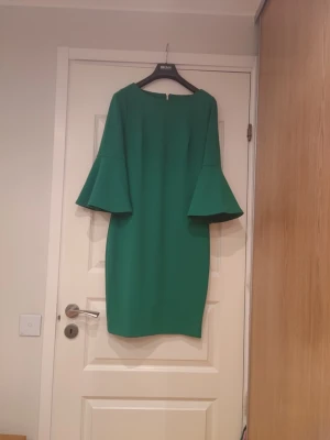 Grön klänning med trumpetärmar - Snygg grön klänning med raka linjer och vida trumpetärmar. Klänningen har en enkel, elegant design och är knälång. Perfekt för dig som gillar stilrena plagg med en twist. Materialet känns slätt och något stadigt.