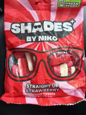 Shades by Niko jordgubbsgodis - Påse med veganskt godis i smaken Straight Up Strawberry från Shades by Niko. Godiset är rött och vitt, format som glasögon. Förpackningen är röd med svart och vit text och innehåller 150 gram sötsaker.