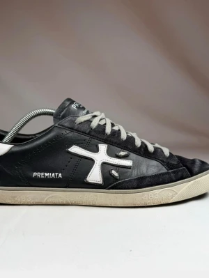 Premiata svarta sneakers med vit detalj - Svarta sneakers från Premiata med vit logotyp på sidan och vita detaljer. Skorna har vita snören och en låg profil. Materialet är en kombination av läder och textil, med en vit gummisula som har mönster och text. Perfekt för dig som gillar stilrena och unika sneakers.