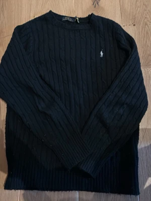 Stickad tröja Ralph lauren - Kabelstickad svart tröja från Polo Ralph Lauren med rund halsringning och broderad vit logga på bröstet. Tröjan har långa ärmar och ribbstickade muddar vid ärmslut och nederkant. Perfekt för en stilren och klassisk look.