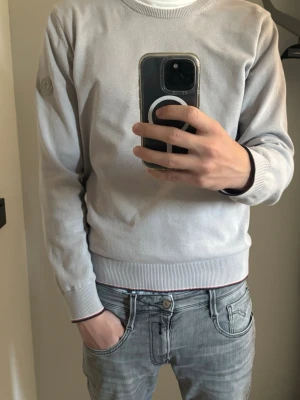 Ljusgrå Moncler sweatshirt - Snygg ljusgrå sweatshirt från Moncler med rund hals och diskret logotyp på vänster ärm. Tröjan har ribbade muddar och nederkant med en tunn röd och marinblå rand. Perfekt för en stilren och avslappnad look.
