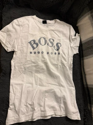 Vit Hugo Boss t-shirt med tryck - Vit t-shirt från Hugo Boss med stort BOSS-tryck i blått framtill. Klassisk rund halsringning och rak passform. Tillverkad i mjuk bomull som känns skön mot huden. Perfekt för en stilren och avslappnad look.