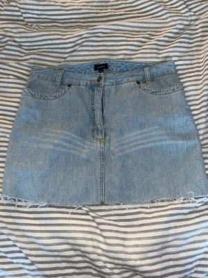 Ljusblå jeanskjol från LINDEX - Säljer en ljusblå jeanskjol från LINDEX med klassisk femficksdesign och rå, fransig fåll. Kjolen har bälteshällor, knapp- och dragkedjestängning samt snygga bakfickor med broderad detalj. Perfekt för en avslappnad och trendig look.