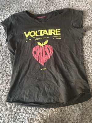 Voltaire svart t-shirt med tryck - Cool svart t-shirt från Zadig & Voltaire med gul text 'VOLTAIRE' och röd 'CRUSH' i form av ett äpple på framsidan. Baksidan har turnélista i gult. Klassisk passform och rund hals, perfekt för en avslappnad look. Se på bilden hur texten ser ut 🫶🏻