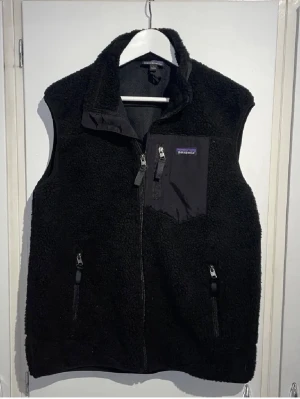 Svart teddyväst från Patagonia - Svart teddyväst från Patagonia med dragkedja framtill, hög krage och tre fickor med dragkedja. Västen har en bröstficka i nylon med logga och är tillverkad i mjukt, fluffigt teddy-material. Perfekt för lager-på-lager och chill stil.