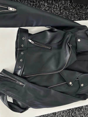 Svart skinnjacka  - Snygg svart skinnjacka med klassisk bikerstil, silvriga dragkedjor och tryckknappar. Jackan har flera fickor med dragkedja och detaljerade sömmar. Perfekt för en cool och trendig look. Den är lite liten i storleken