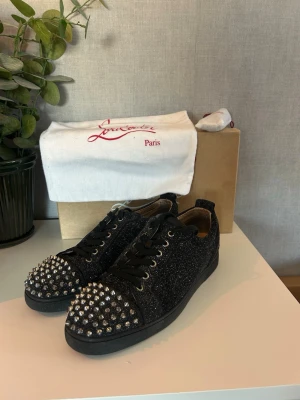 Svarta glittriga Louboutin sneakers - Svarta sneakers från Christian Louboutin med glittrig ovandel och silverfärgade nitar över tån. Skorna har klassisk låg modell, svarta skosnören och detaljer i läder. Perfekta för dig som vill sticka ut med en edgy och lyxig stil. allt og