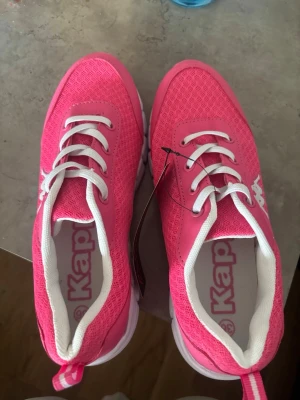 Rosa sneakers från Kappa - Snygga rosa sneakers från Kappa med vit sula och vita detaljer. Ovandelen är i meshmaterial för bra andningsförmåga och skorna har klassisk snörning. Perfekta för dig som vill sticka ut med färg på fötterna.