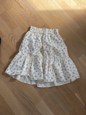 Vit blommig volangkjol - Supersöt vit kjol med små rosa blommor och volangkant. Kjolen har resår i midjan och är gjord i ett lätt bomullstyg, perfekt för varma dagar. Den har en luftig och romantisk känsla som passar till många olika stilar.