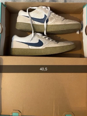 Nike vita och blå sneakers - Säljer ett par vita Nike sneakers med blå swoosh och beige mockadetaljer. Skorna har lågt skaft och en grönaktig gummisula. Perfekta för dig som gillar klassisk streetstil och vill ha något som sticker ut lite extra.