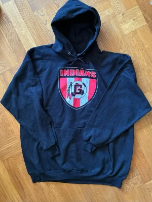 Svart vintage hoodie med Indians-logga US - Svart hoodie med stor röd och vit Indians-logga på bröstet och huva med dragsko. Klassisk känguruficka framtill och ribbade muddar. Perfekt för dig som gillar sportig och avslappnad stil. Size XXL