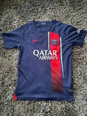 PSG matchtröja Nike blå/röd - Snygg Paris Saint-Germain matchtröja från Nike i mörkblått med röd och vit rand på framsidan. PSG-logga på bröstet, Qatar Airways-tryck och korta ärmar. Perfekt för fotbollsträning eller att bära som supporter. Materialet är lätt och andas bra.