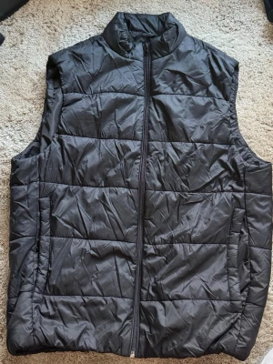 Svart dunväst från ZARA - Snygg svart dunväst från ZARA med hög krage och dragkedja framtill. Västen har en quiltad design och två sidofickor. Perfekt att slänga över en hoodie eller tröja för en cool lager-på-lager-look.  Aldrig använd