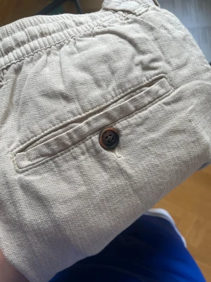 Beiga linnebyxor med resårmidja - Snygga beiga linnebyxor med resår i midjan och två bakfickor med knappar. Perfekta för en avslappnad och stilren look. Byxorna har rak passform och är tillverkade i ett luftigt linnematerial.