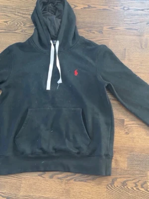 Svart hoodie från Polo Ralph Lauren - Svart hoodie från Polo Ralph Lauren med klassisk röd broderad logga på bröstet. Hoodien har vit dragsko, stor känguruficka och mjukt material. Perfekt för en avslappnad och stilren look.