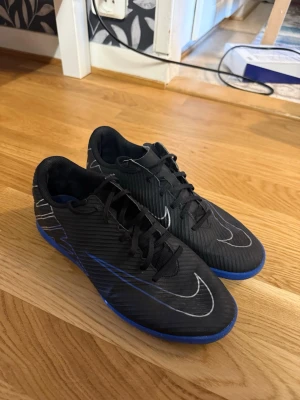 Svarta Nike fotbollsskor med blå sula - Snygga svarta fotbollsskor från Nike med blå sula och vita detaljer. Skorna har klassisk snörning och en låg profil, perfekt för snabba rörelser på planen. Materialet är syntetiskt och designen har en modern look med stora Nike-loggor på sidan. Storlek 40 och har andvänts bara inomhus!