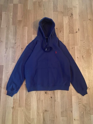 Mörkblå Ralph Lauren Zip hoodie - Storlek M - Snygg marinblå hoodie från Polo Ralph Lauren med röd broderad logga på bröstet. Tröjan har huva med snörning, dragkedja framtill och två stora fickor. Tillverkad i mjuk bomull som är skön att ha på sig. Perfekt för chill dagar och streetwear. Skick enligt bilder.