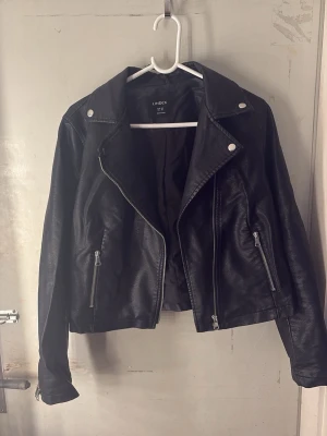 Svart skinnjacka  - Snygg svart skinnjacka från Lindex med klassisk bikerstil. Jackan har silvriga dragkedjor, bred krage och tryckknappar på axlarna. Två fickor framtill med dragkedja och en cool, tidlös look som passar till det mesta. Saken man hänger upp jackan på är sönder men det är ju inget som förändrar dess utseende utom den är bara jobbigare att hänga upp !