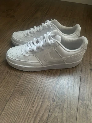 Nike Court Vision Low - Klassiska helvita Nike Air Force 1 sneakers i lågt utförande. Skorna har perforerad tå för ventilation, vit sula och snörning. Tillverkade i läder med ikoniska swoosh-loggan på sidan. Perfekta för en clean och stilren look.
