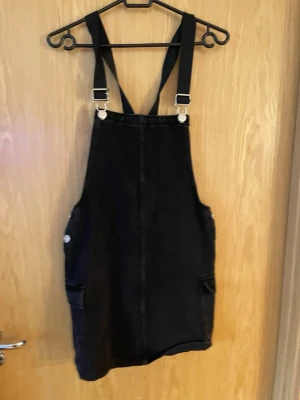 Svart denim overall kjol från C&A - Cool svart denim overall kjol från C&A med justerbara axelband och fickor på sidorna. Klassisk modell med knappar i metall. Perfekt att styla med en t-shirt eller hoodie under för en avslappnad look.