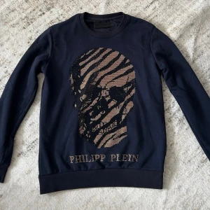 Svart sweatshirt med dödskalle - Svart sweatshirt med stort glittrigt dödskalletryck i guld och svart. Tröjan har rund halsringning och ribbade muddar vid ärmslut och nederkant. Perfekt för dig som gillar statement-plagg.
