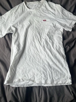 Vit t-shirt från Levi's med logga - Klassisk vit t-shirt från Levi's med liten röd logga på bröstet. T-shirten har rund halsringning och korta ärmar. Tillverkad i mjuk bomull för en skön och avslappnad stil.