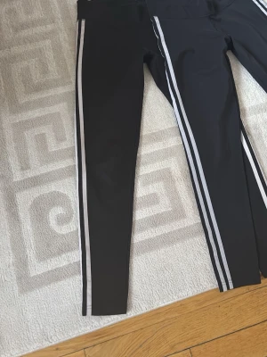 Svarta leggings med vita ränder - Snygga svarta leggings med två vita ränder längs sidorna. Tight passform och hög midja, perfekta för en sportig och trendig look. Materialet är stretchigt och bekvämt, vilket gör dem lätta att röra sig i hela dagen. Vi säljer både två 