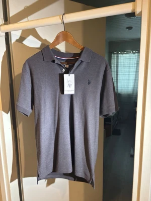 Grå pikétröja från U.S Polo Assn. - Klassisk grå pikétröja från U.S Polo  med korta ärmar och krage. Tröjan har knappslå framtill och den ikoniska broderade loggan på bröstet. Perfekt för en stilren och avslappnad look.
