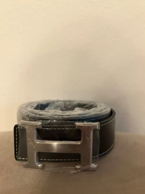  svart skärp med H-spänne - Svart skärp från med ikoniskt H-format spänne i silverfärgad metall. Skärpet har vita kontrastsömmar längs kanterna och är tillverkat i skinn. Perfekt accessoar för att lyfta din outfit.
