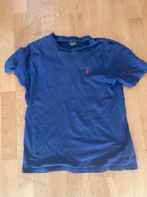 Blå t-shirt från Polo Ralph Lauren - Snygg marinblå t-shirt från Polo Ralph Lauren med klassisk röd logga broderad på bröstet. T-shirten har rund hals och korta ärmar, perfekt för en avslappnad och stilren look. Tillverkad i mjuk bomull för extra komfort.