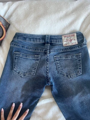 Blå True Religion jeans med brodyr - Säljer ett par klassiska blå jeans från True Religion med deras ikoniska broderade bakfickor och patch i midjan. Jeansen har en rak passform och är tillverkade i denim med snygga detaljer. Perfekta för en trendig och avslappnad stil.