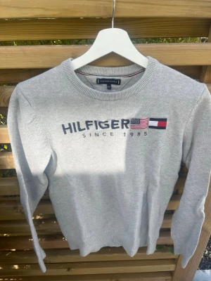 Grå tröja Tommy Hilfiger - Snygg grå långärmad tröja från Tommy Hilfiger med broderad logga och flagga framtill. Tröjan har rund halsringning och ribbade muddar vid ärmslut och nederkant. Perfekt för dig som gillar klassisk och stilren design.