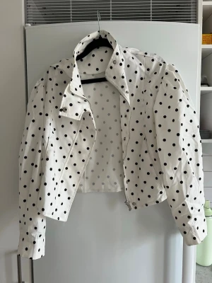 Vit prickig kort jacka - Snygg vit jacka med svarta prickar och croppad passform. Jackan har dragkedja framtill, hög krage och långa ärmar. Perfekt för dig som vill sticka ut med ett lekfullt mönster.