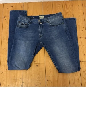 Blå Morris  Steve Satin Jeans med brodyr - Säljer ett par klassiska blå jeans från Steve Satin Jeans. Modellen har fem fickor, snygga slitningar och en broderad detalj på myntfickan. Jeansen är raka i modellen och tillverkade i mjukt denimtyg. Perfekta till vardagslooken!