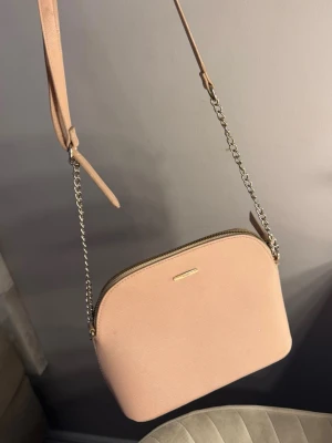Ljusrosa axelväska från Michael Kors - Snygg ljusrosa axelväska från Michael Kors med stilren design. Väskan har justerbar axelrem i kombination av kedja och läder, samt guldfärgade detaljer och dragkedja. Perfekt för att bära det viktigaste med stil.