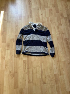 Randig rugbytröja från Ralph Lauren - Snygg långärmad rugbytröja från Ralph Lauren med breda marinblå och beige ränder, vit krage och knappslå. Stor siffra 3 på ryggen och liten logga på bröstet.  Storlek är S (8) 